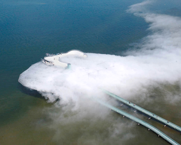 arial shot of fog-covered architectural pavilion suspended over a lake, with a long bridge walkway leading into a cloud-like structure.