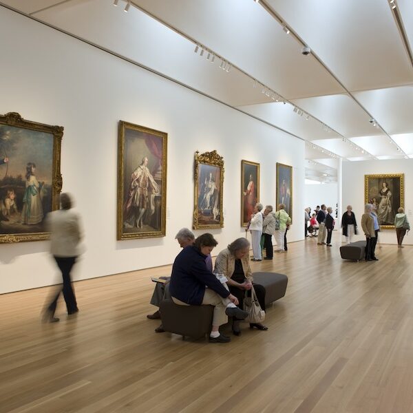 A spacious art gallery with people admiring classic paintings displayed on white walls, and wooden flooring, creating a warm atmosphere.