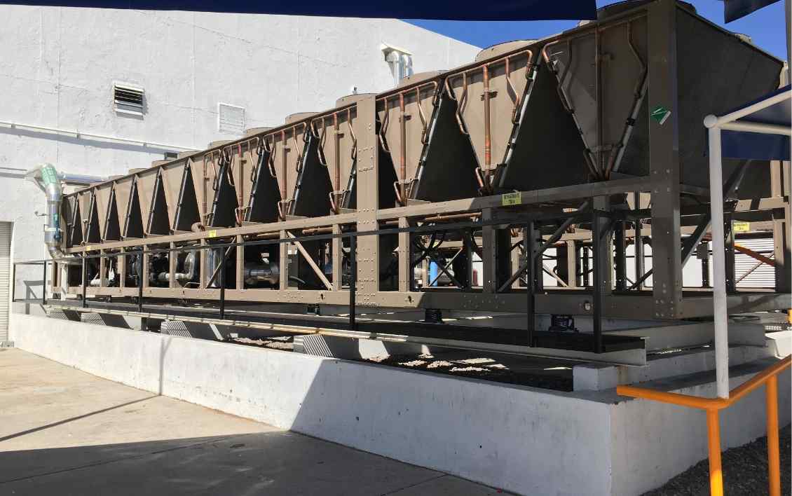 A long, industrial cooling unit with multiple connected compartments, mounted on a platform against a plain building wall.
