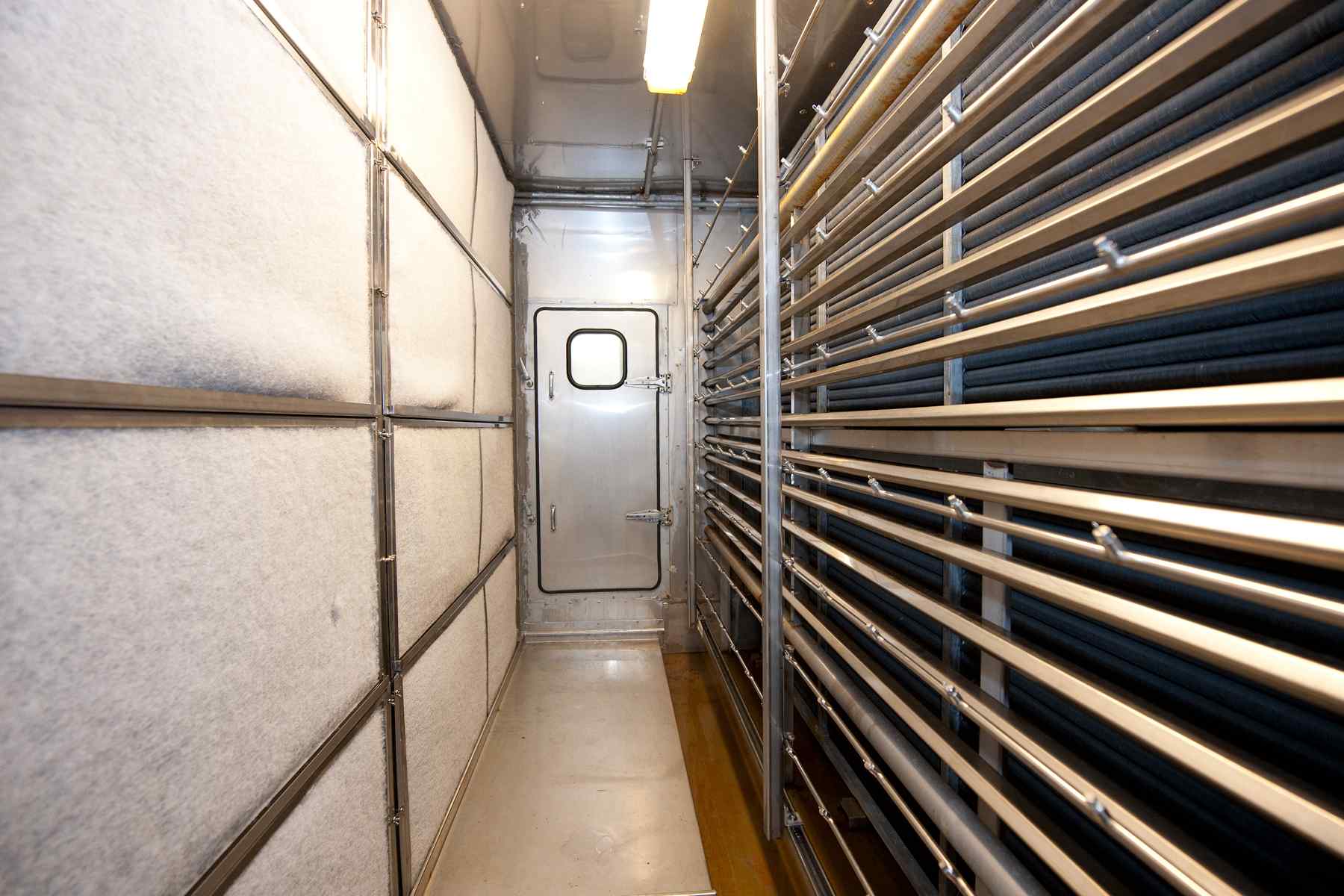 Interior of a transport vehicle, featuring padded walls, metal rods, and a door, illuminated by overhead lighting.