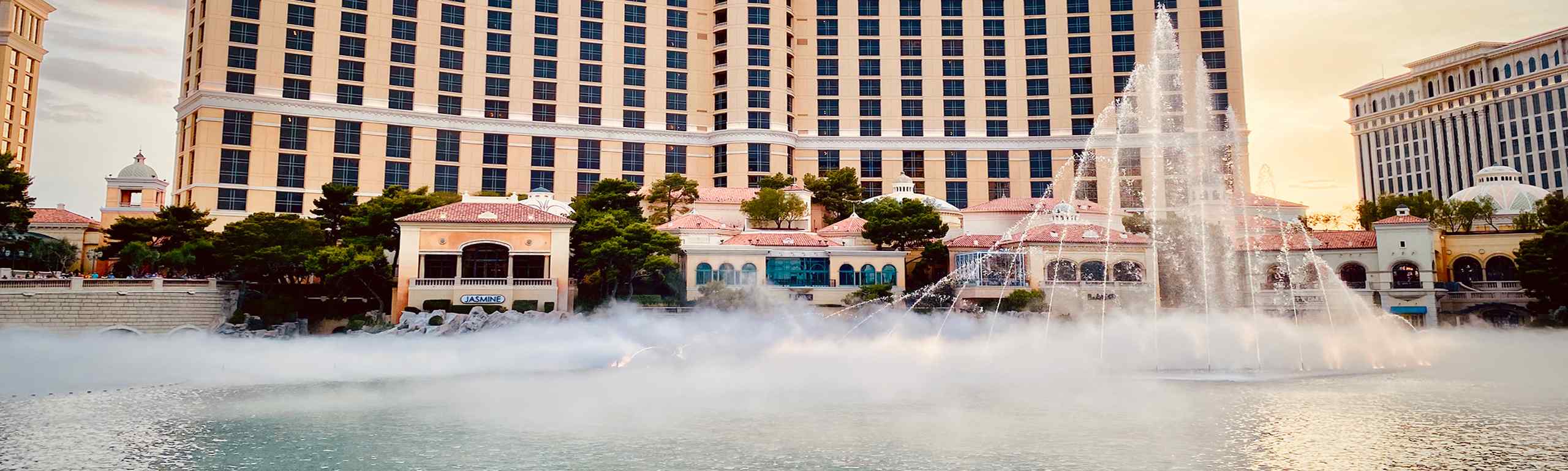 A view of the Bellagio Hotel's iconic fountains with mist, surrounded by lush greenery and elegant architecture at sunset.