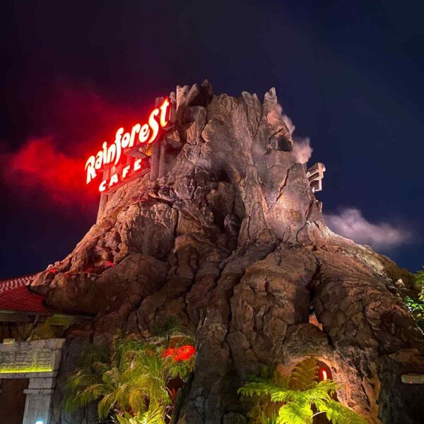 The Rainforest Cafe sign glows atop a towering artificial mountain, surrounded by lush greenery, under a dark night sky.