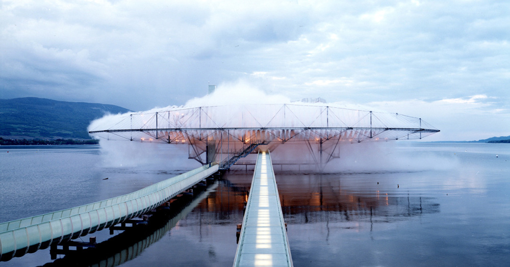 A fog-covered architectural pavilion suspended over a lake, with a long bridge walkway leading into a cloud-like structure—the Blur Building at Swiss Expo 2002.