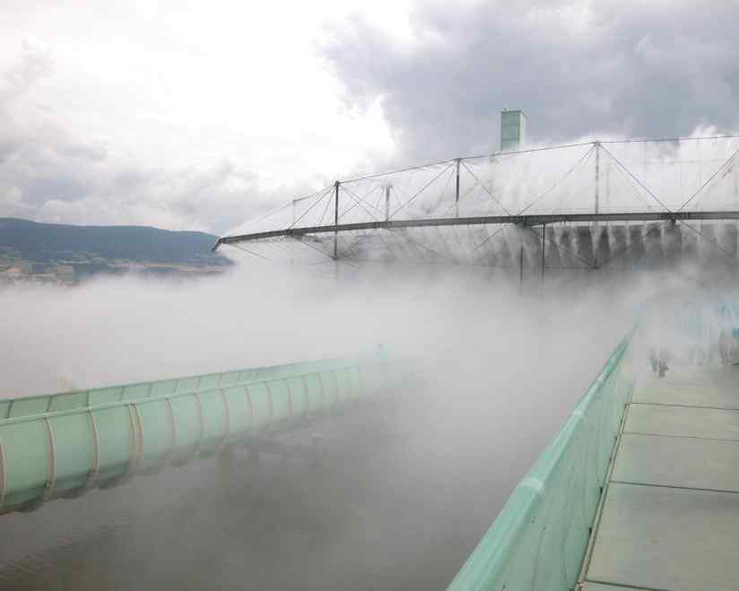 A misty landscape featuring a long water slide, with a large structure above dispersing fog against a cloudy sky.