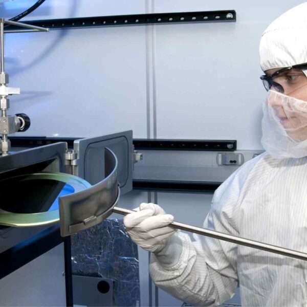 A technician in a protective suit operates machinery with a tube, handling a circular, transparent object inside a cleanroom environment.