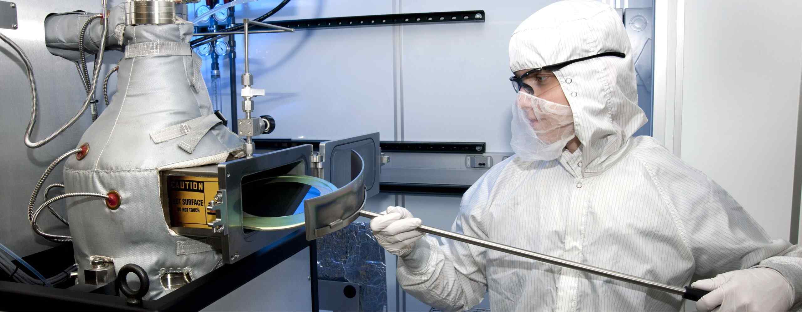 A technician in a protective suit operates machinery with a tube, handling a circular, transparent object inside a cleanroom environment.