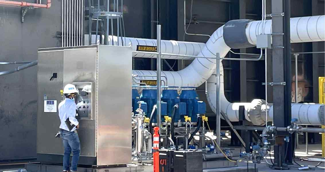 A worker in a hard hat inspects equipment in an industrial facility, surrounded by large pipes and machinery.