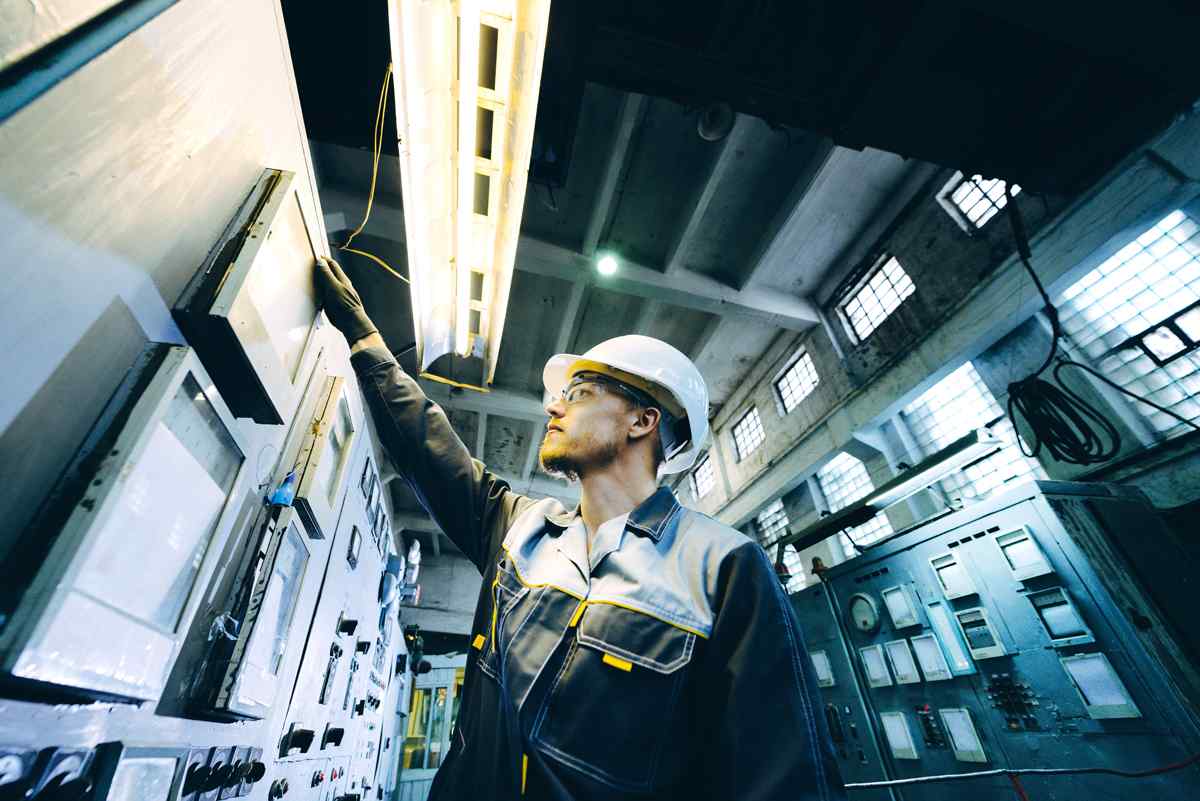An industrial worker in a hard hat adjusts equipment on a control panel inside a well-lit facility with large windows.