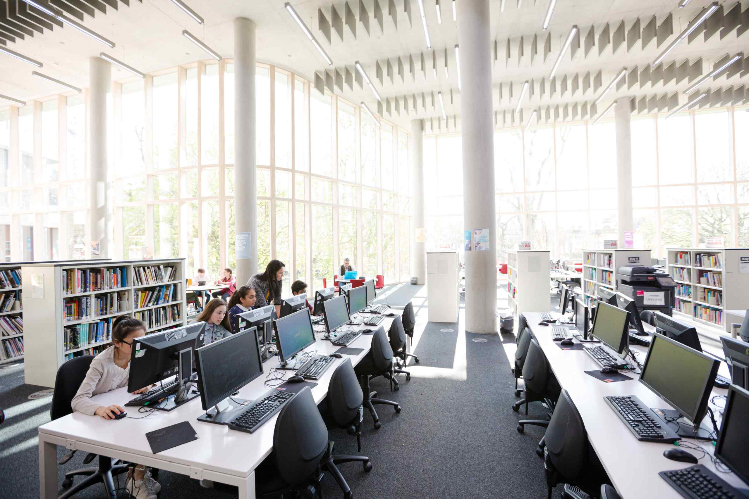 A bright library interior featuring computer stations, bookshelves, and large windows allowing natural light to fill the space.