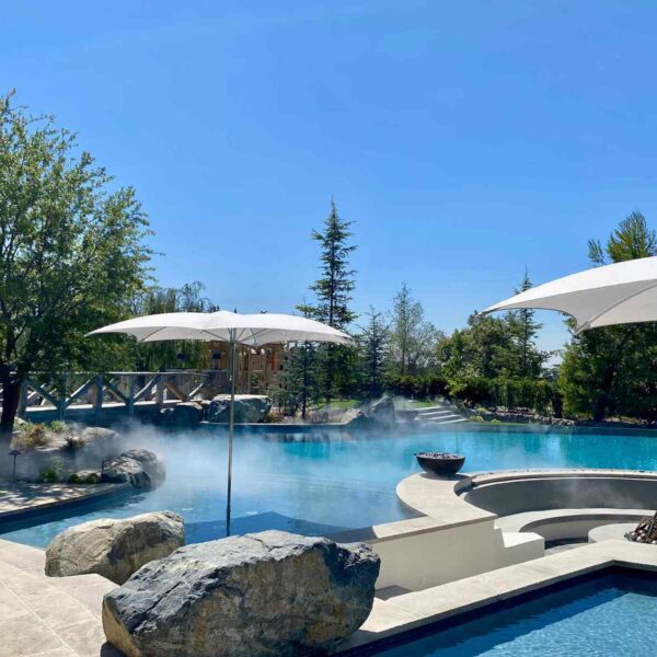 A serene pool scene featuring mist, large rocks, white umbrellas, and lush greenery under a clear blue sky.