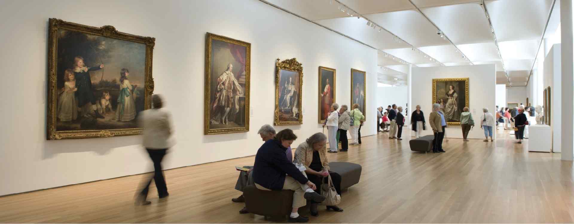 A spacious art gallery filled with visitors admiring large, framed paintings on white walls, enjoying the elegant interior design.