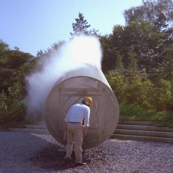 A worker in a hard hat inspects a large metallic cylinder emitting steam, surrounded by greenery and gravel path.