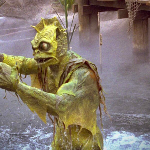 A green, aquatic creature stands in murky water, reaching out with clawed hands, surrounded by fog and vegetation.