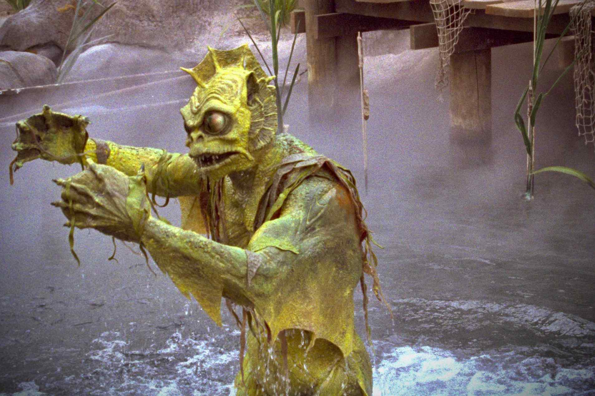 A green, aquatic creature stands in murky water, reaching out with clawed hands, surrounded by fog and vegetation.