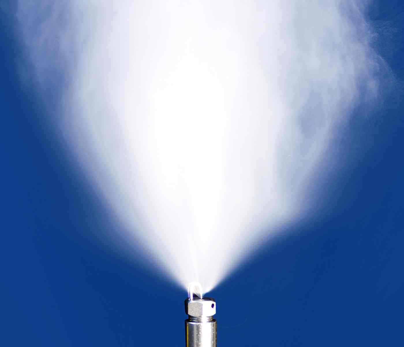 Close-up of a nozzle emitting a fine mist against a bright blue background, creating a cloud-like spray.
