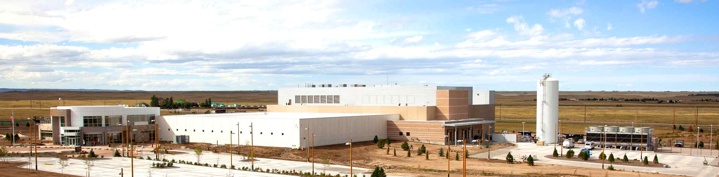 A modern industrial facility with a large main building and storage silos, set against a vast landscape under a cloudy sky.