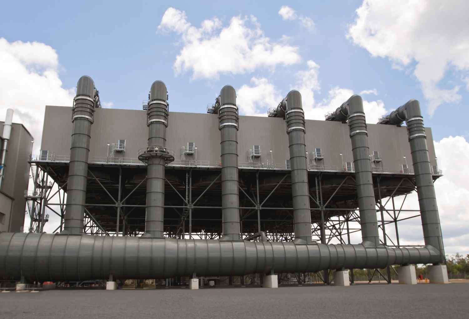 Industrial facility with large metallic pipes extending from a high structure, under a cloudy sky, indicating energy production or processing.