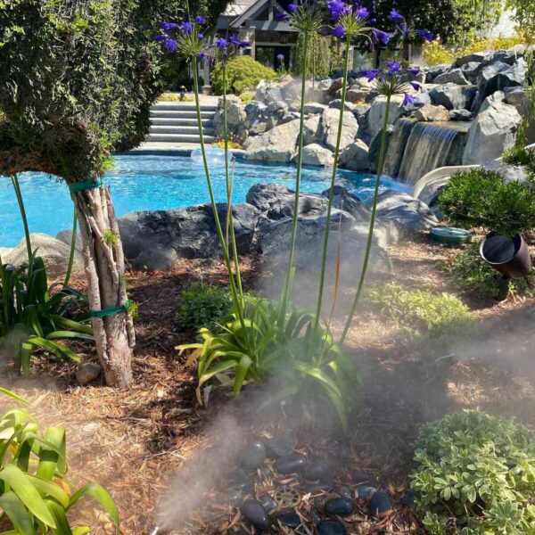 Lush garden featuring tall purple flowers, a swimming pool, a waterfall, and mist rising from the ground amidst greenery and stones.