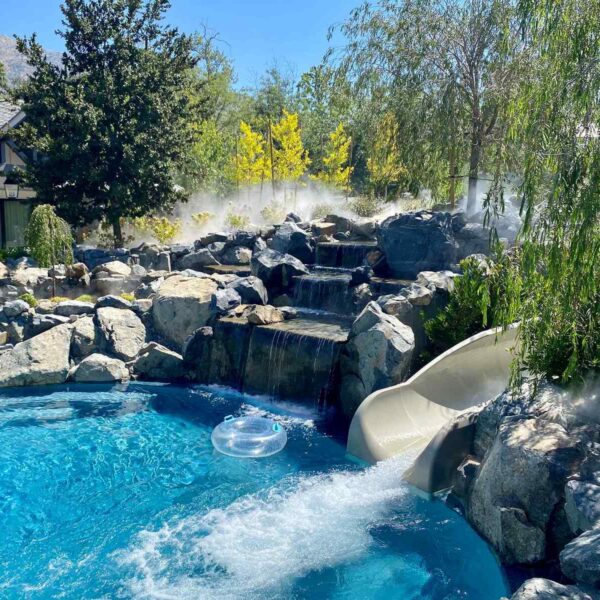 A luxurious backyard pool features a waterfall, a water slide, and a floating inner tube, surrounded by lush greenery and mist.