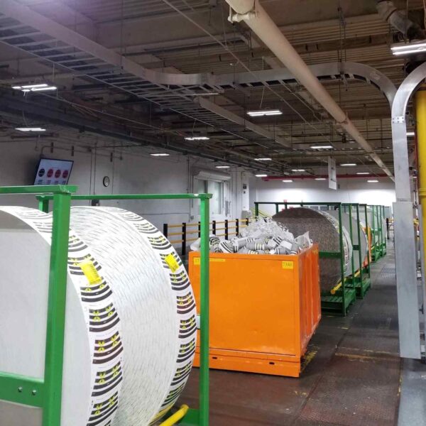 Image of a warehouse interior featuring large rolls of paper on green carts alongside orange containers, under bright overhead lighting.