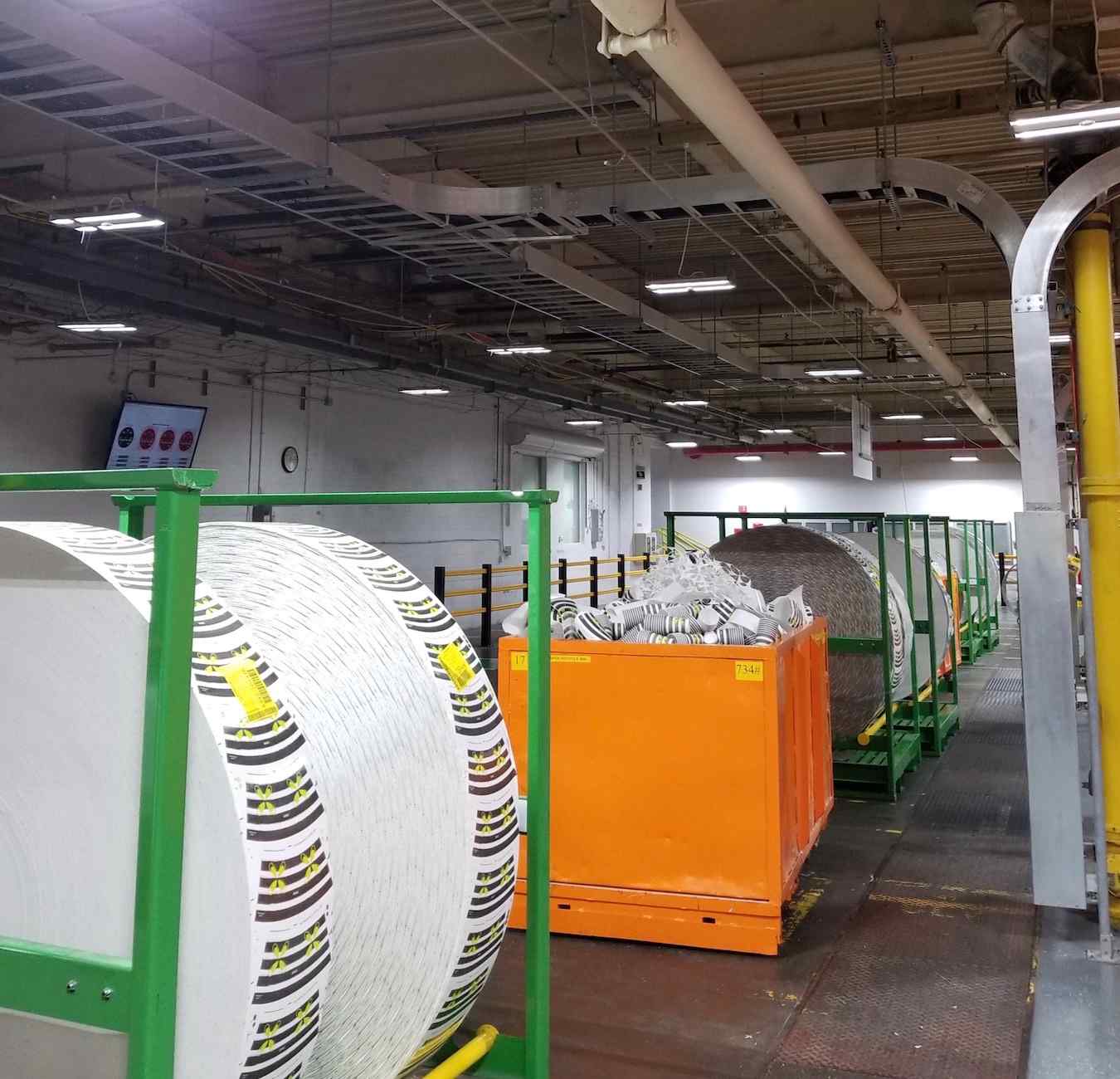 Image of a warehouse interior featuring large rolls of paper on green carts alongside orange containers, under bright overhead lighting.
