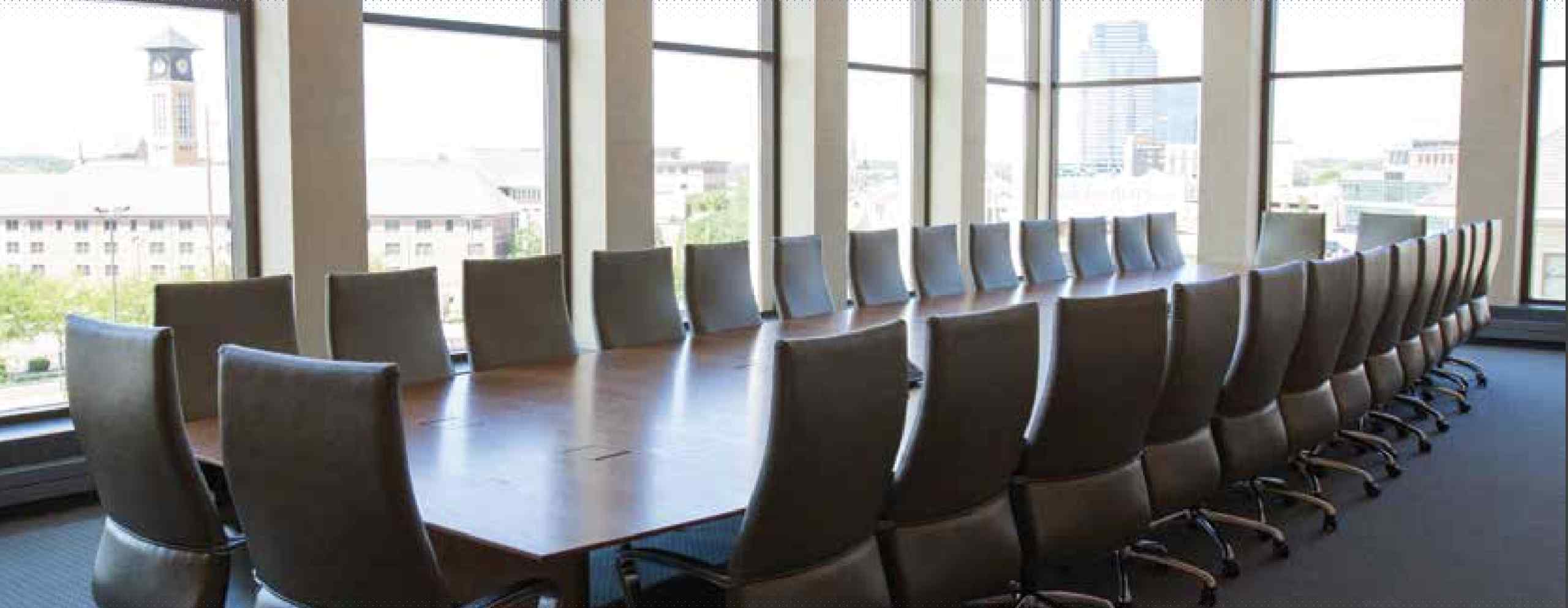 A spacious conference room with a long wooden table and numerous leather office chairs, surrounded by large windows showing a city view.
