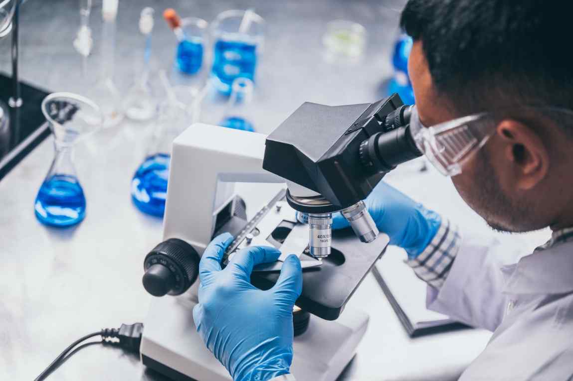 A scientist in gloves examines a sample under a microscope in a lab filled with blue liquids in beakers and flasks.