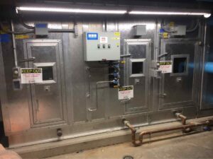 Metallic enclosure with two access doors, warning signs to keep out, and control panel, set against a concrete wall.