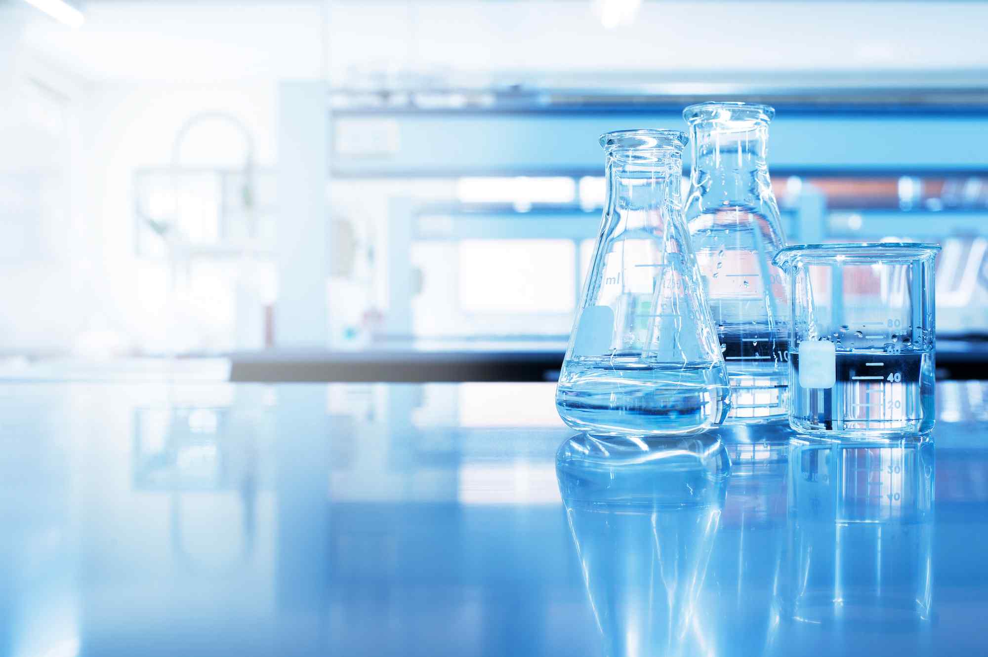 Three clear glass laboratory vessels filled with liquid, reflecting on a glossy surface in a bright, modern lab environment.