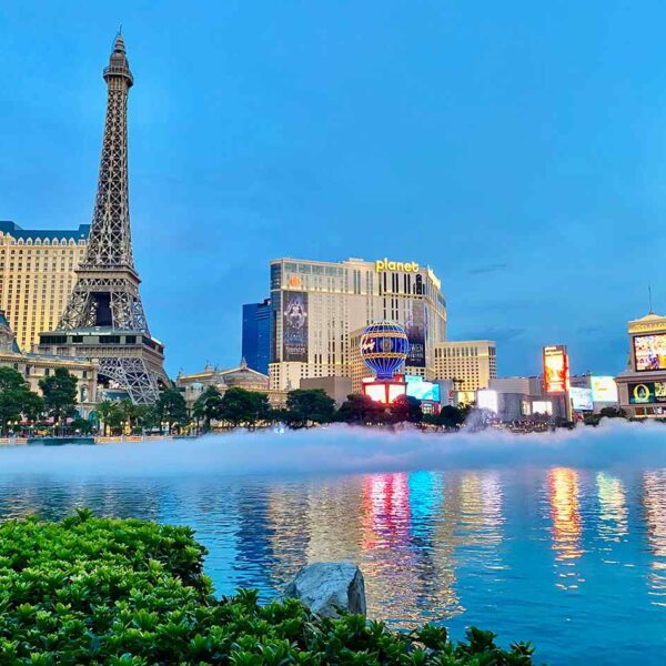 Las Vegas skyline at dusk featuring the Eiffel Tower replica, vibrant billboards, and shimmering reflections in the water.