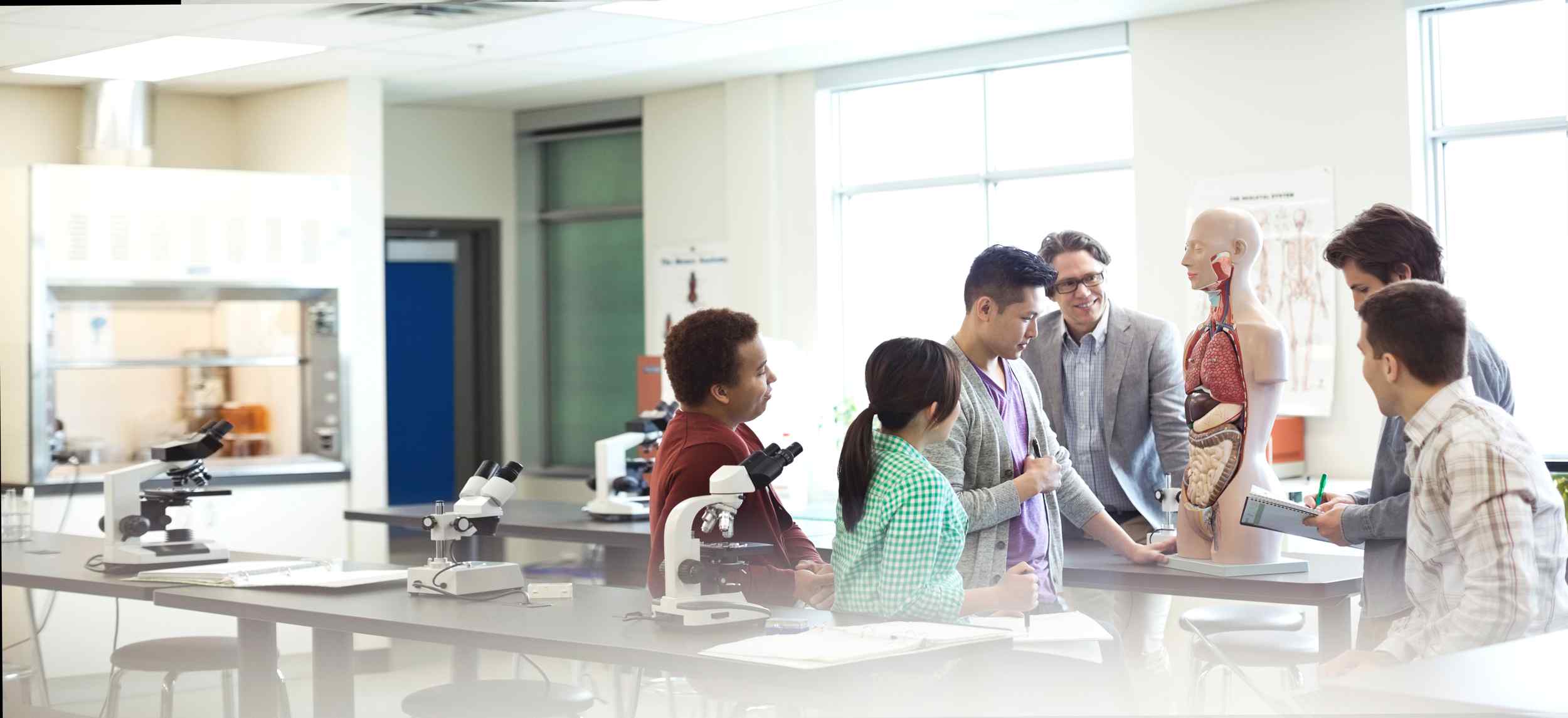 A diverse group of students engages in discussion around an anatomical model in a bright laboratory setting. Microscopes are visible on the tables.