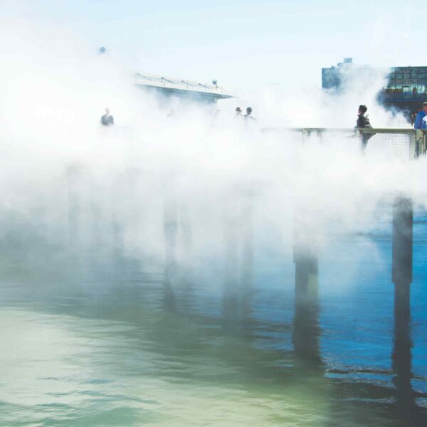 A foggy pier extends over calm water, with people walking in the mist, creating a serene and ethereal atmosphere under blue skies.
