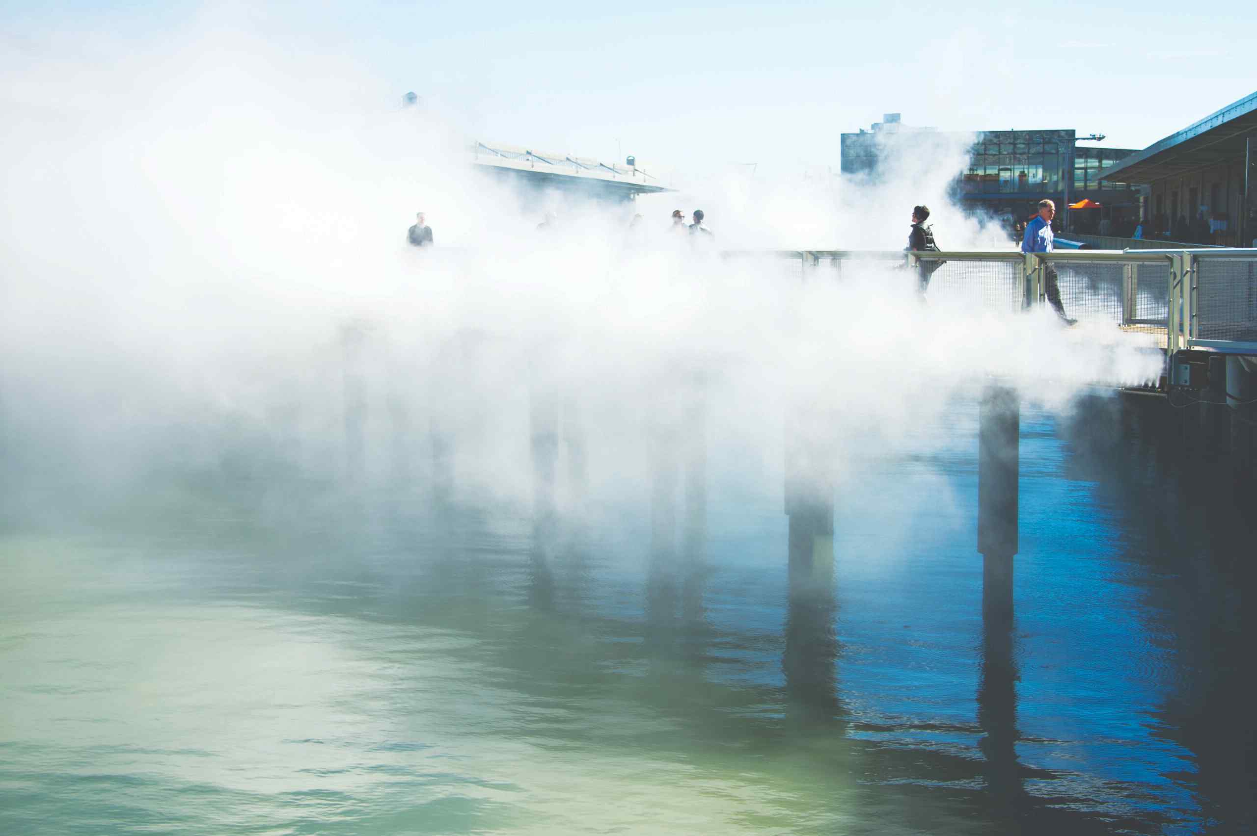 A foggy pier extends over calm water, with people walking in the mist, creating a serene and ethereal atmosphere under blue skies.