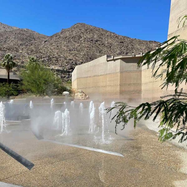 A serene outdoor space features water fountains amidst mist, set against a backdrop of rocky mountains and palm trees under a clear blue sky.