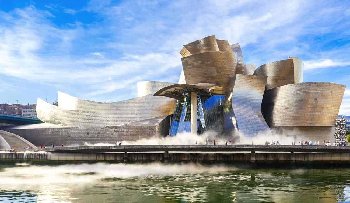 Guggenheim Museum Bilbao's striking, curvilinear facade reflects sunlight as it overlooks the river, with visitors strolling nearby.