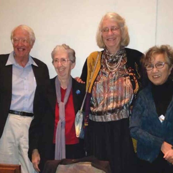 A group of four adults stands together, dressed in a variety of colorful outfits, smiling and posing for the photo indoors.