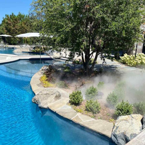 A luxurious swimming pool surrounded by lush landscaping, with gentle mist rising from the garden, under a clear blue sky.