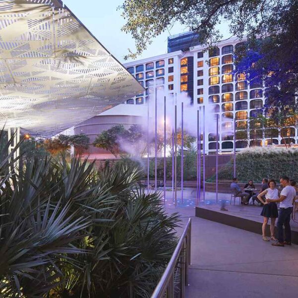 Couple stands together in a modern outdoor space with decorative canopy, fog elements, lush greenery, and vibrant seating areas.