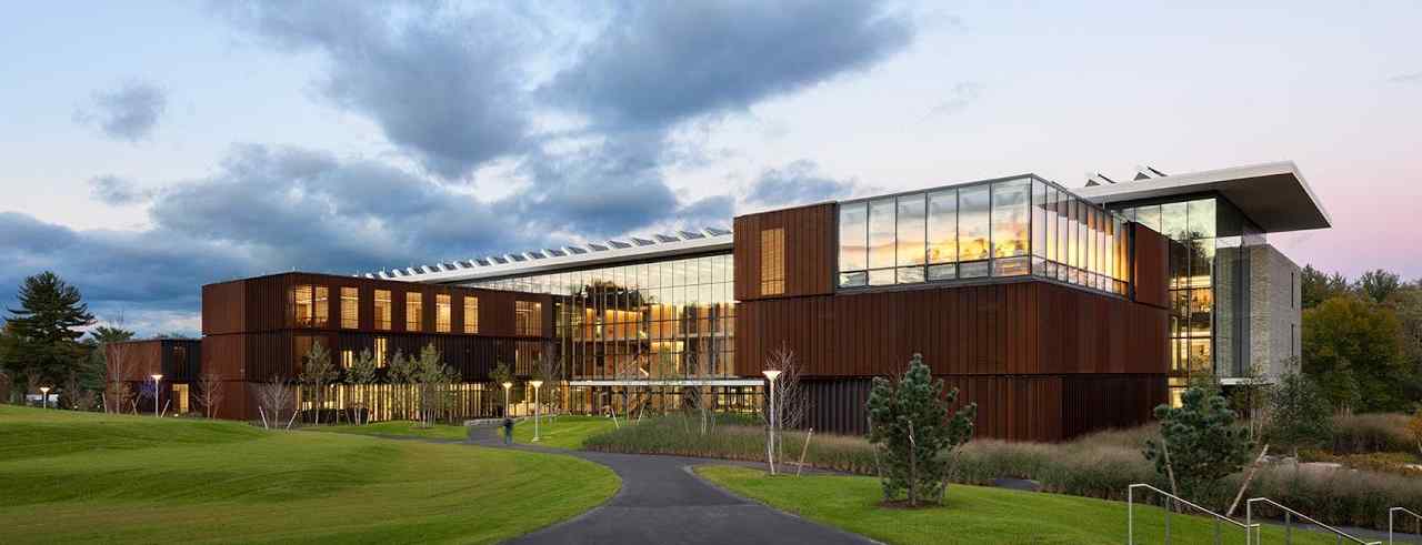 Modern building with a mix of glass and metal, surrounded by lush green lawns and trees under a serene twilight sky.