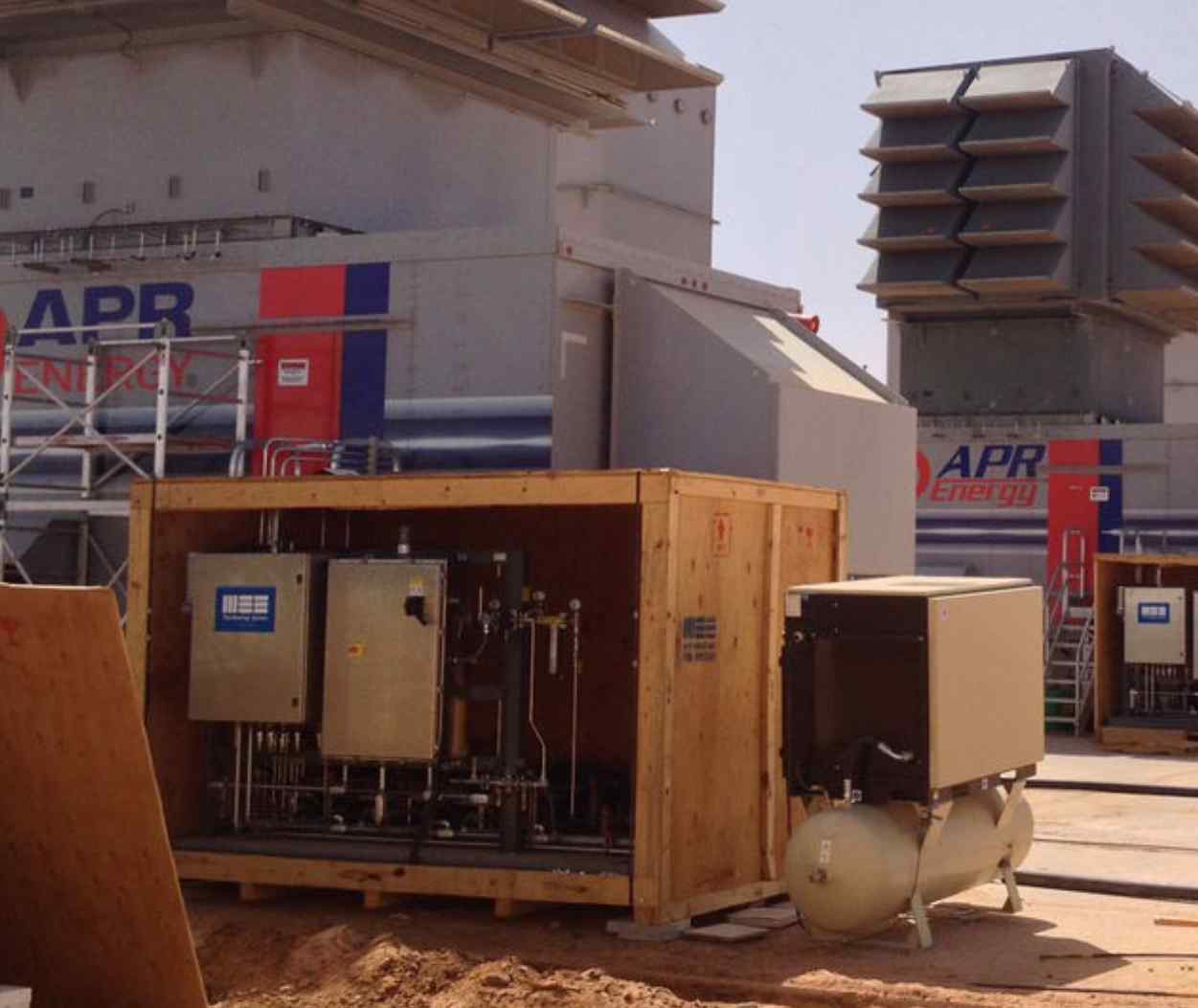 An industrial setup featuring APR Energy equipment, including a wooden enclosure with machinery and large cooling units in the background.