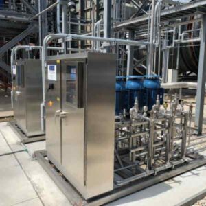 Industrial equipment featuring two stainless steel control cabinets and blue pumps connected by numerous pipes in a processing facility.