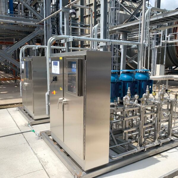 Industrial equipment featuring two stainless steel control cabinets and blue pumps connected by numerous pipes in a processing facility.