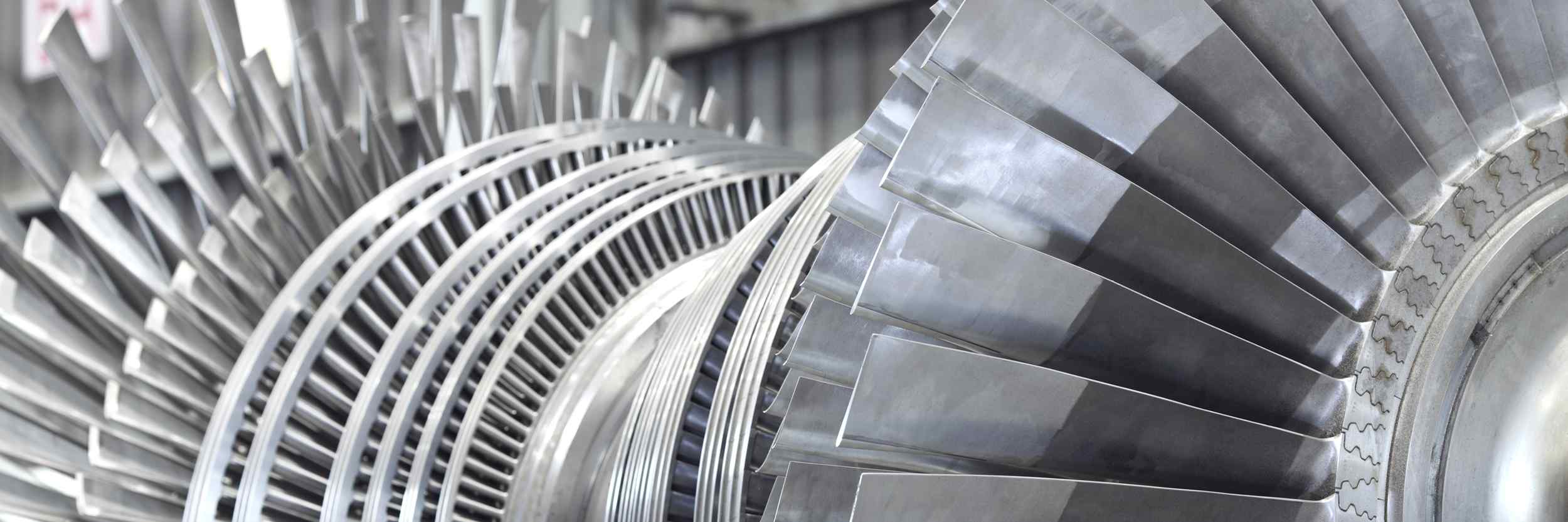 Close-up of metallic turbine blades, showcasing their detailed design and structure, with a blurred industrial background.