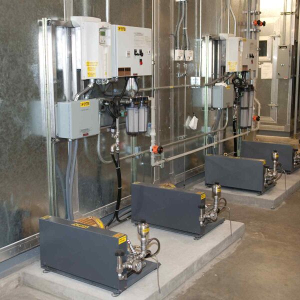 Three industrial pumps connected to control panels and piping on a concrete floor, set against a metallic wall in a facility.