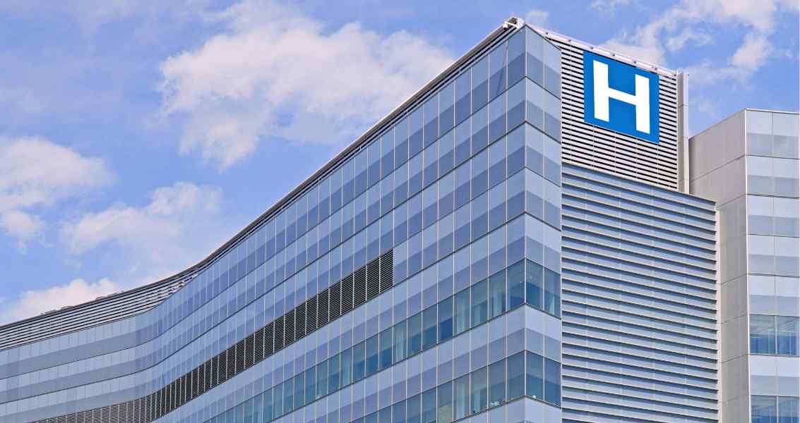 Modern hospital building with glass facade, featuring a prominent blue hospital sign against a bright sky.