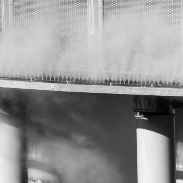 Black and white image of a mist emitting from a metallic structure, supported by large, shadowed columns.