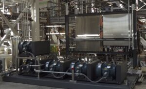 Industrial assembly area featuring multiple machinery motors connected by pipes, with scaffolding and stainless steel surfaces in the background.