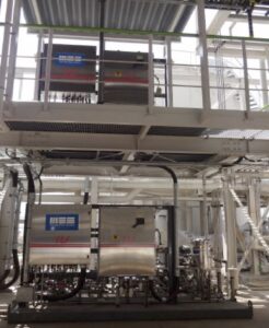 Industrial equipment setup with two metal control panels, pipes, and a walkway, located in a bright, spacious facility.