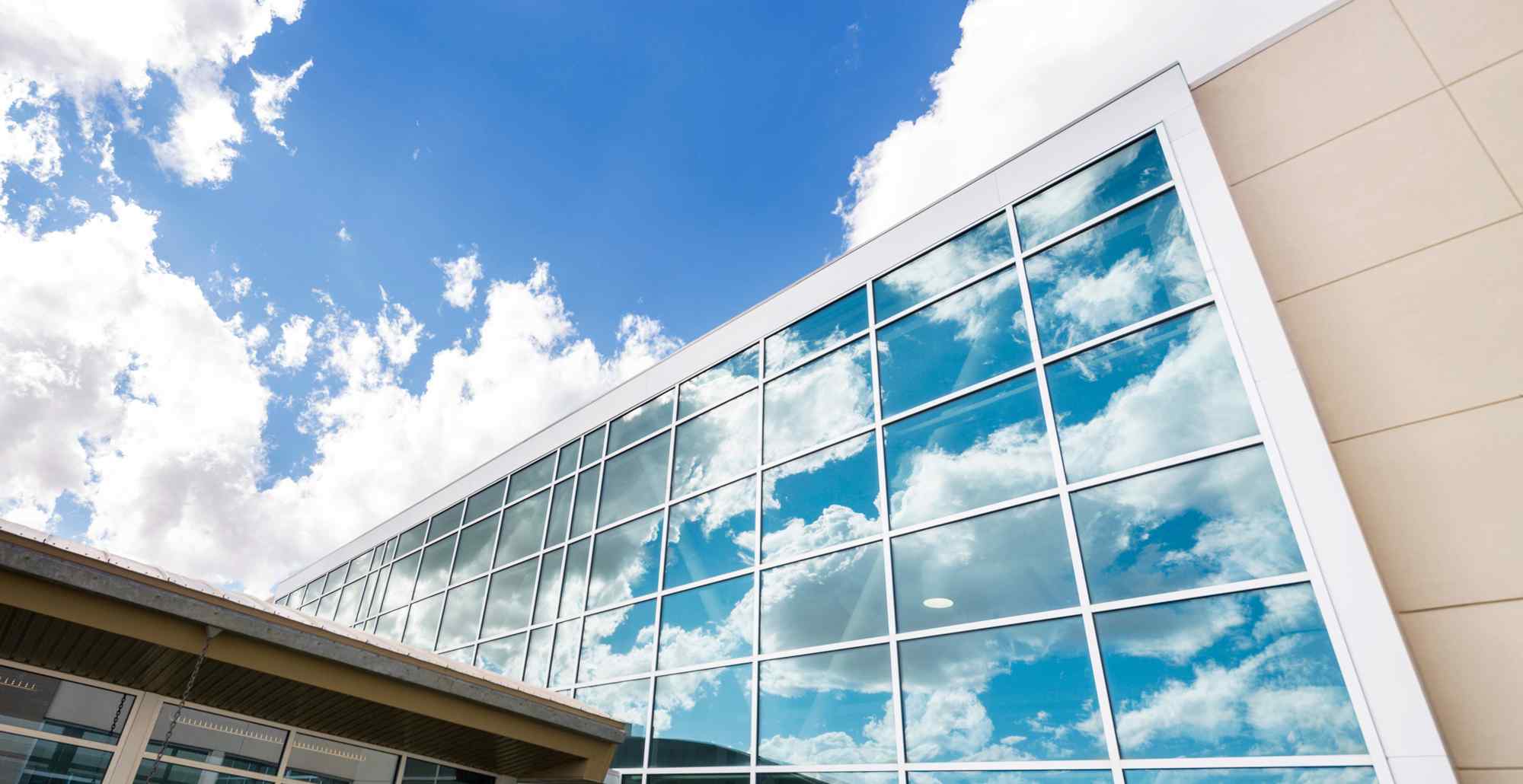A modern building with large glass windows reflecting a bright blue sky filled with fluffy white clouds.