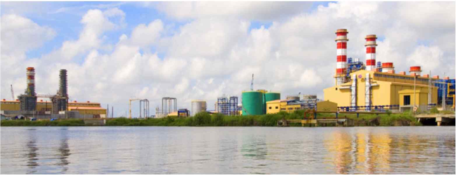 Industrial power plant with large chimneys and storage tanks, reflected in calm water under a partly cloudy sky.